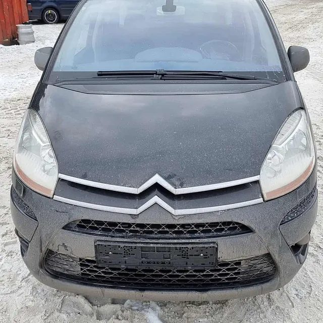 Grå Citroën varebil parkert på snødekt område foran verksted og stablede biler.