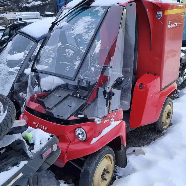 Rød elektrisk arbeidsbil med lukket førerhus parkert i snødekt område ved verksted.