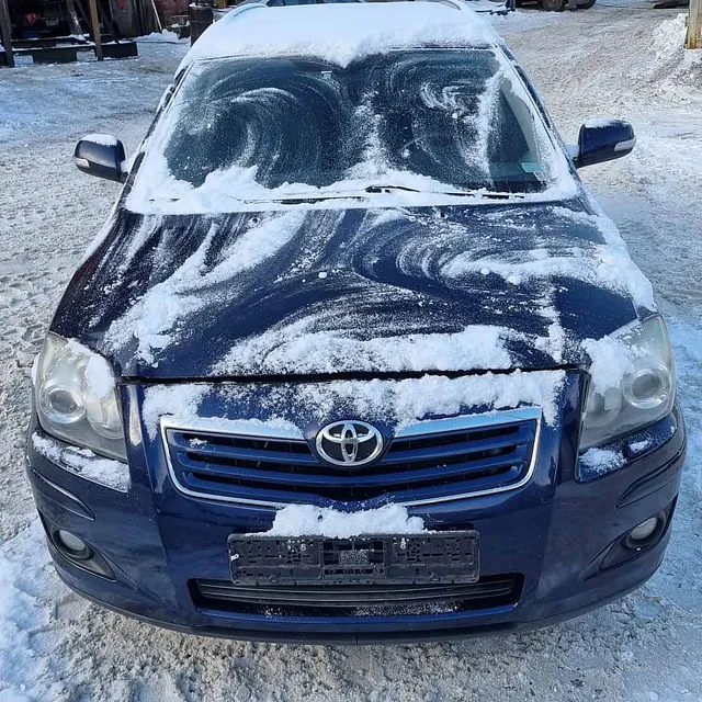 Blå personbil dekket av snø parkert på bilopphuggeri med verksted og utstyr i bakgrunnen.