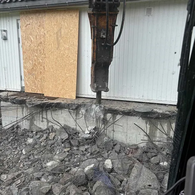 Hydraulisk pigghammer knuser betongfundament ved bygg under rivningsarbeid.