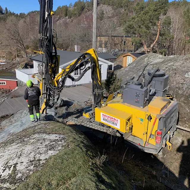 Borerigg utfører fjellboring på tomt med boligområde og skog i bakgrunnen.