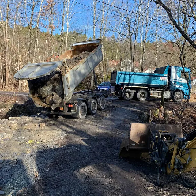 Lastebil tipper stein og masser fra henger under anleggsarbeid på grusvei i skogsområde.
