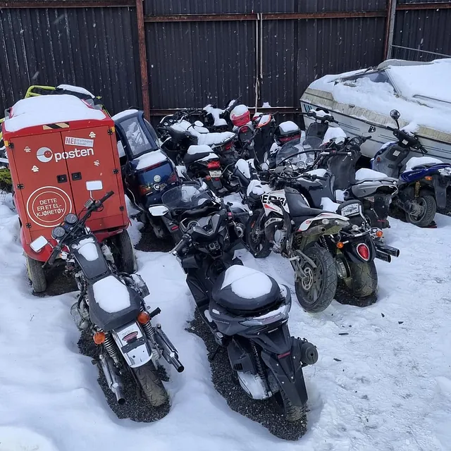 Flere motorsykler parkert tett sammen på snødekt område bak gjerde i industriområde.