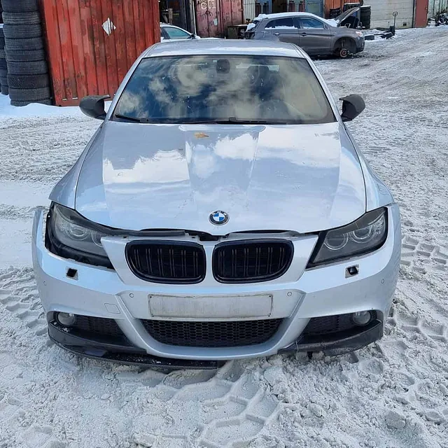 Hvit BMW med snø på panser parkert på bilopphuggeri med containere og biler bak.