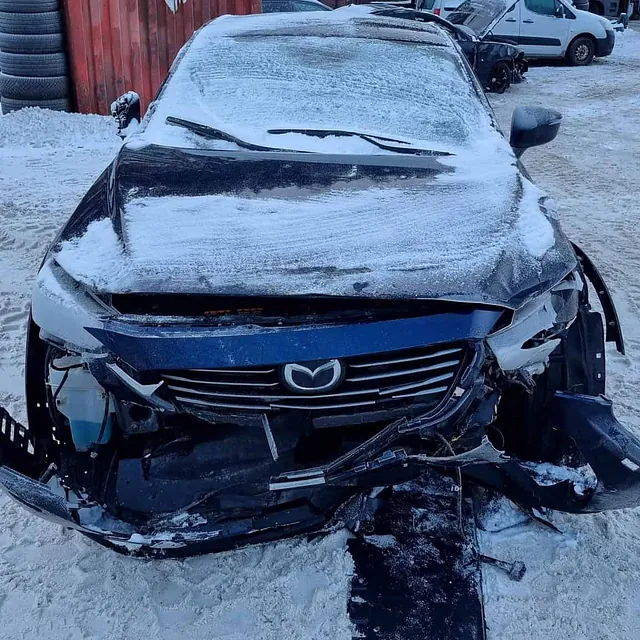 Blå Mazda med kraftig frontskade og snø på panser parkert på opphuggeri.