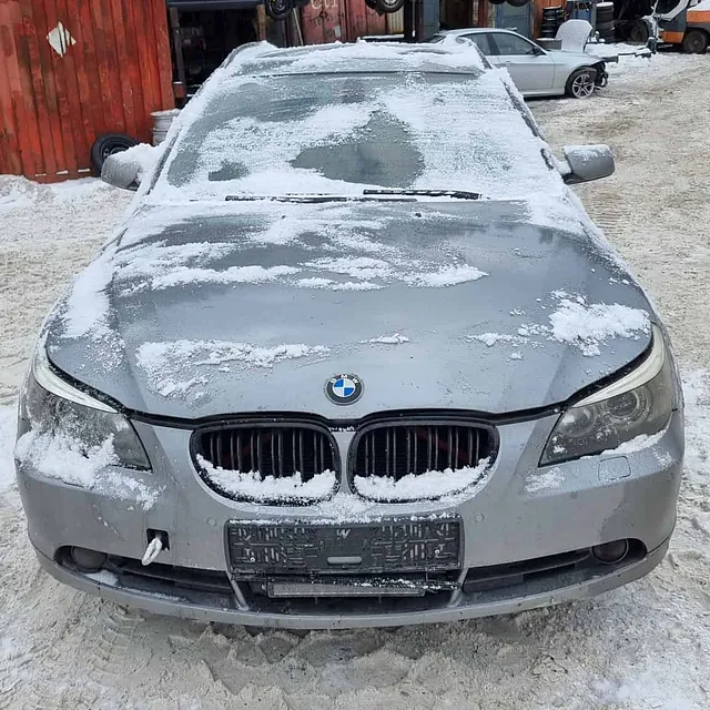 Sølv BMW med snø på panser parkert på snødekt område ved verksted og biler bak.