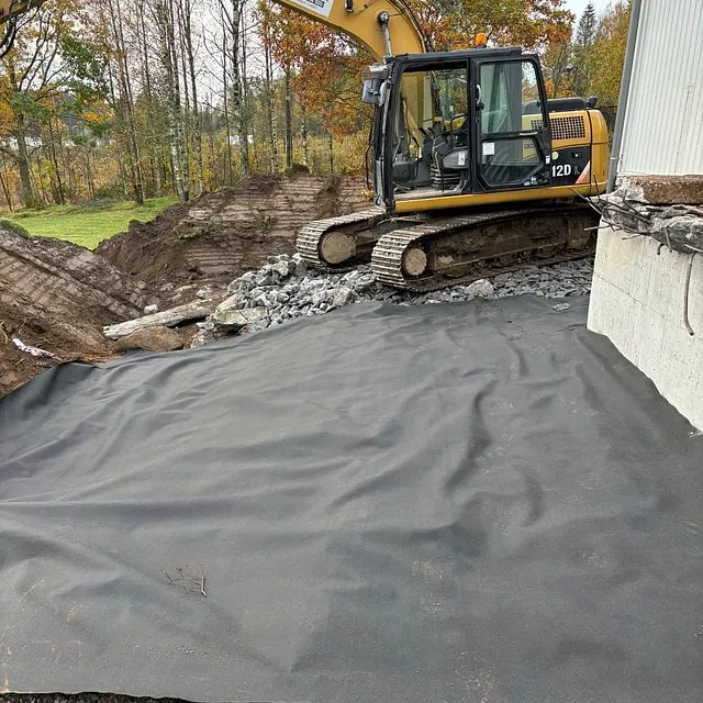 Gravemaskin utfører grunnarbeid og klargjør tomt med duk og pukk ved bolig.