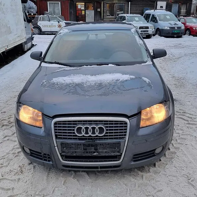Grå Audi med lysene på parkert på snødekt område med bygninger og biler bak.